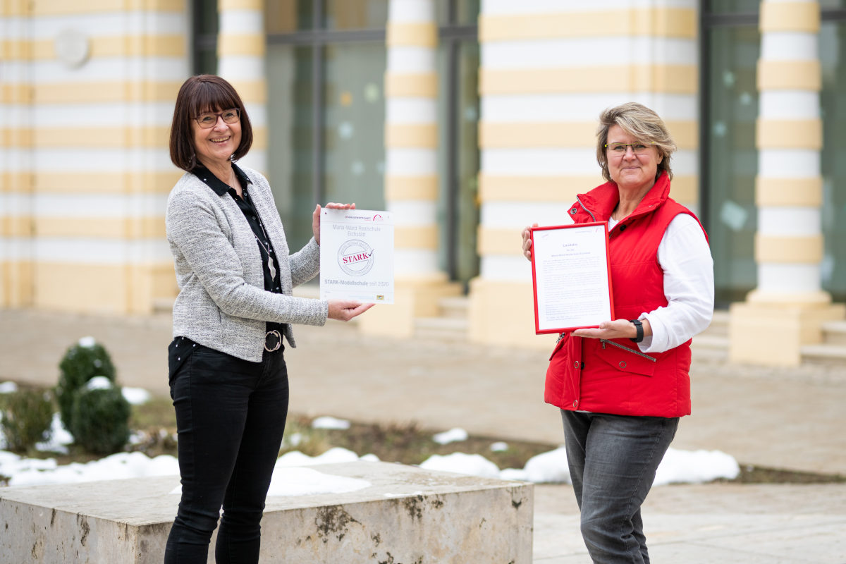 Zwei Vertreterinnen der Schule stehen mit Abstand auf dem Schulgelände der Maria‑Ward‑Realschule Eichstätt und halten eine gerahmte Laudatio sowie das STARK‑Gütesiegel in die Kamera.