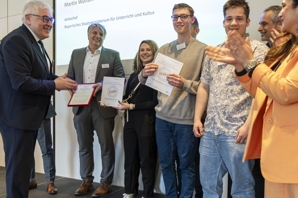 Mehrere Schüler*innen und Lehrkräfte der ausgezeichneten Münchner Berufsschulen für das Bäcker-/Konditoren- und Metzgerhandwerk stehen gemeinsam mit Schulleiter Theo Albrecht auf einer Bühne und freuen sich über den Erhalt der STARK-Gütesiegel; links Dieter Vierlbeck.