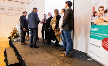 Vertreter*innen der FOS Neu‑Ulm stehen auf der Bühne, während Sven Meyer‑Huppmann vom Bayerischen Staatsministerium für Unterricht und Kultus und Hubert Schurkus, Präsident der bbw‑Gruppe, das STARK‑Gütesiegel und die gerahmte Laudatio überreichen und den Geehrten gratulierend die Hände schütteln.