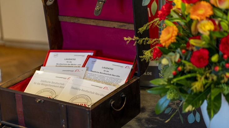 Geöffnete Truhe mit gerahmten Laudationes und Acryl‑Gütesiegeln neben einem Blumenbouquet