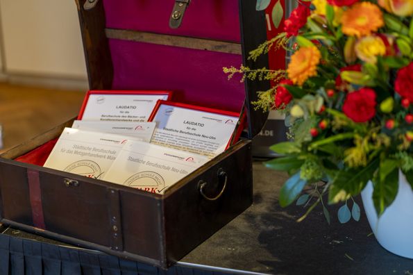 Geöffnete Truhe mit gerahmten Laudationes und Acryl‑Gütesiegeln neben einem Blumenbouquet