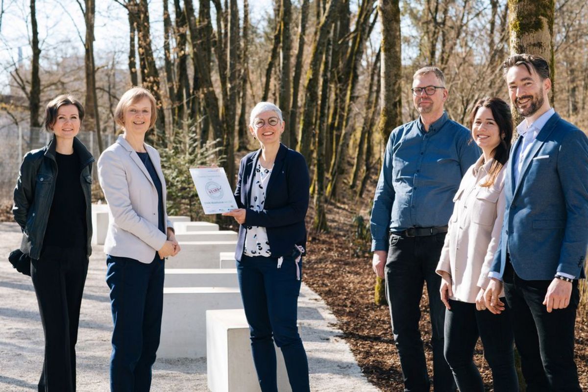 Fünf Lehrkräfte und die STARK‑Projektleiterin im herbstlichen Schulhof der FOS Haar; ein Mann hält eine gerahmte Laudatio, eine Frau ein Acryl‑Gütesiegel zur Auszeichnung als STARK‑Modellschule 2021