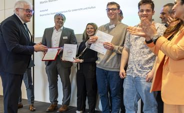 Mehrere Schüler*innen und Lehrkräfte der ausgezeichneten Münchner Berufsschulen für das Bäcker-/Konditoren- und Metzgerhandwerk stehen gemeinsam mit Schulleiter Theo Albrecht auf einer Bühne und freuen sich über den Erhalt der STARK-Gütesiegel; links Dieter Vierlbeck